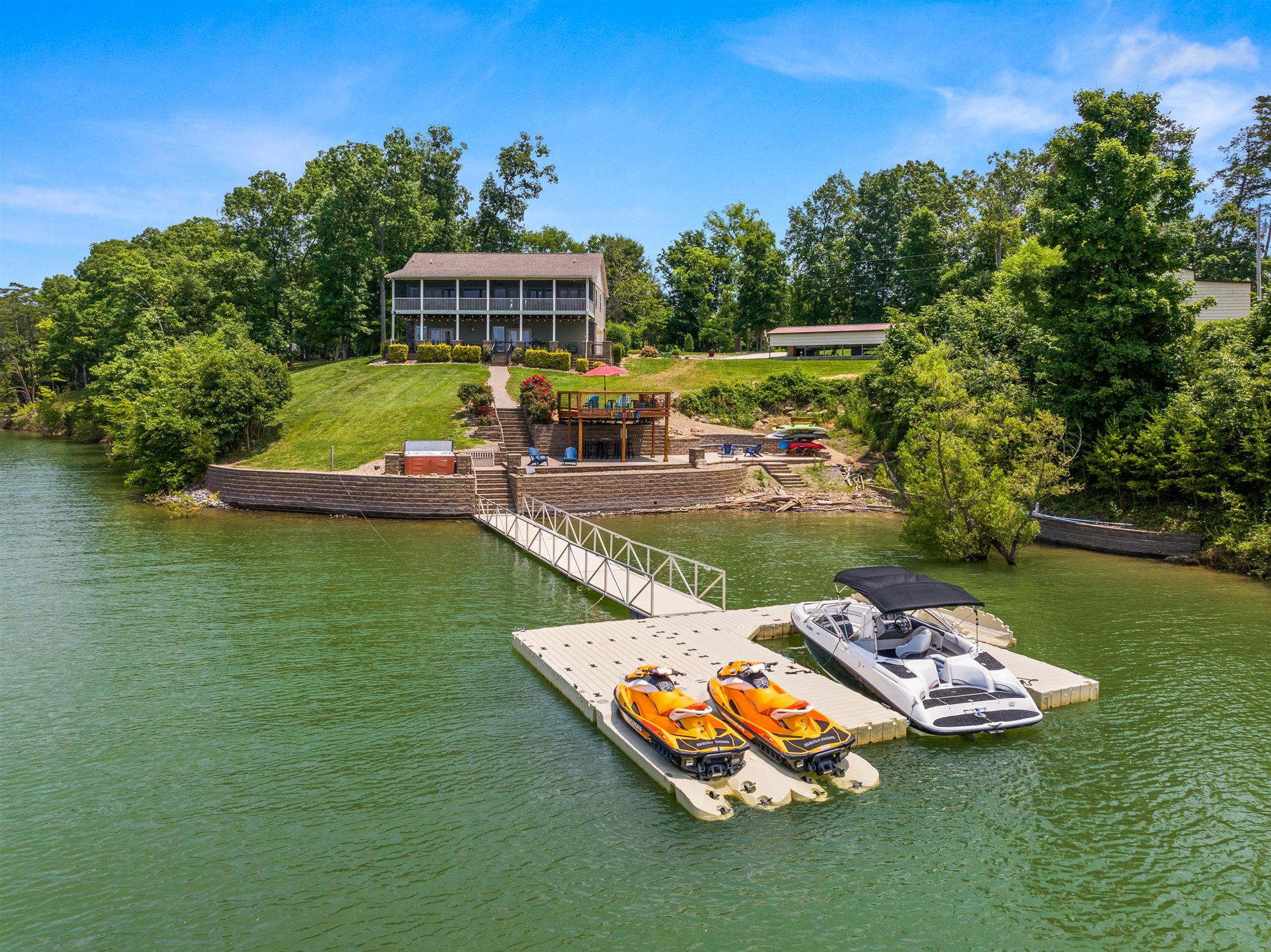 Aerial view from the lake showing the two-story house with wraparound porches, stone patio area, hot tub, and floating dock with two jet skis and a boat