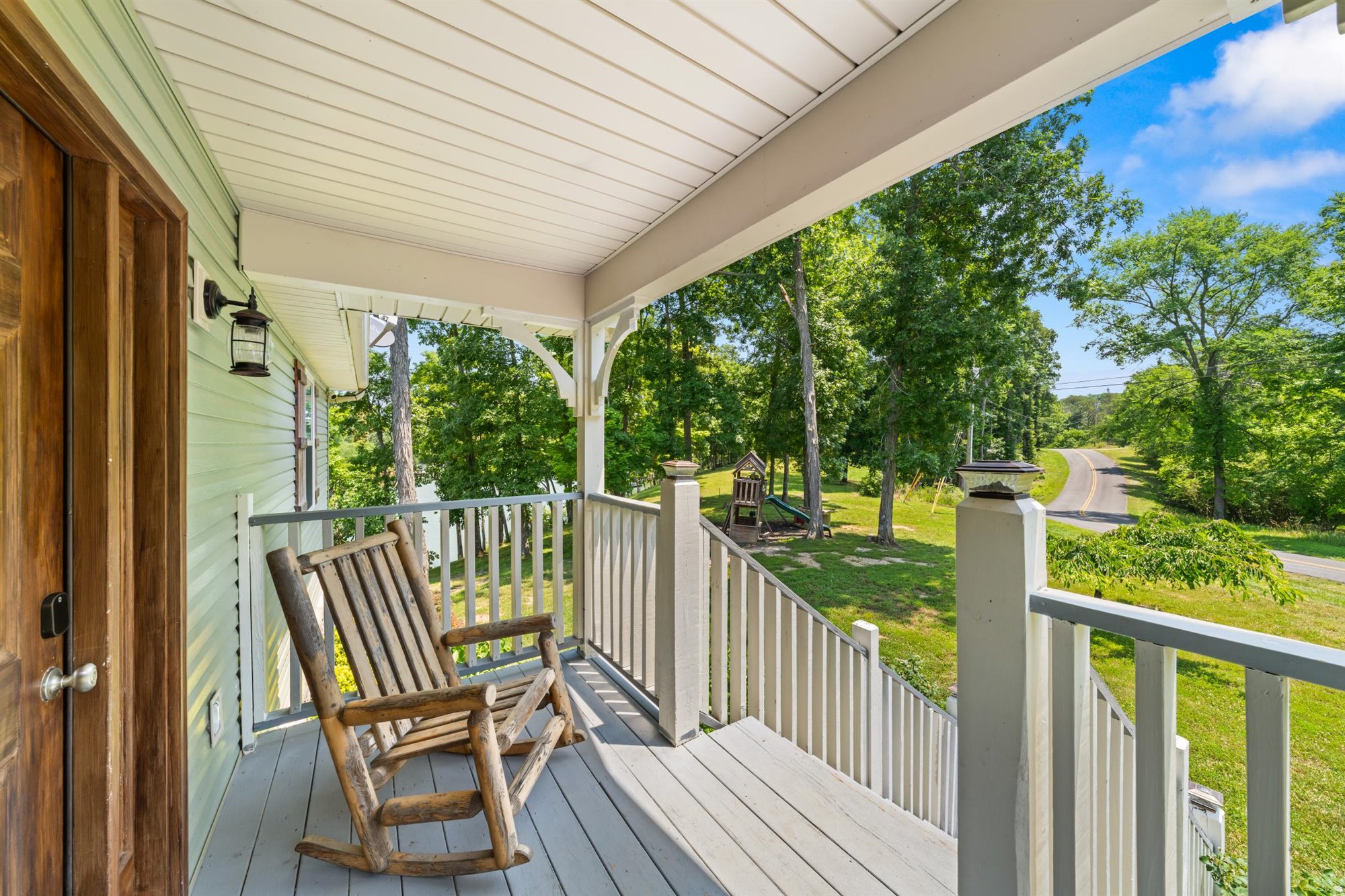 Covered front porch with wooden rocking chair, white railings, and view of the tree-lined driveway and green lawn