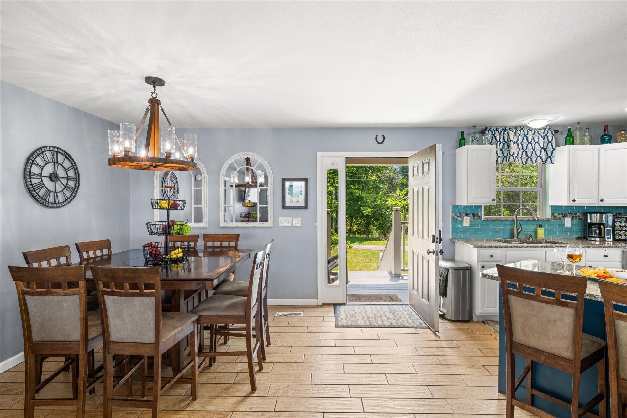 Open concept dining area and kitchen with rustic wagon wheel chandelier, high-top dining table seating six, blue accent walls, white cabinets with turquoise subway tile backsplash, and french doors to the front porch